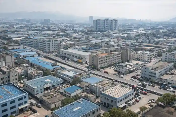 Aerial view of a bustling urban industrial area with clustered buildings, blue-roofed warehouses, narrow roads with vehicles, and hazy mountains in the distant background.