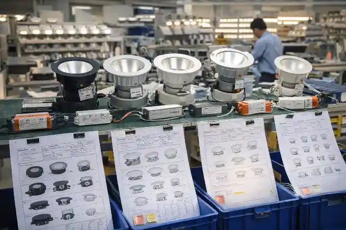Factory workbench displaying black, silver, and white spotlights and downlights with wires, components, blueprints, and blue bins in a manufacturing setting.