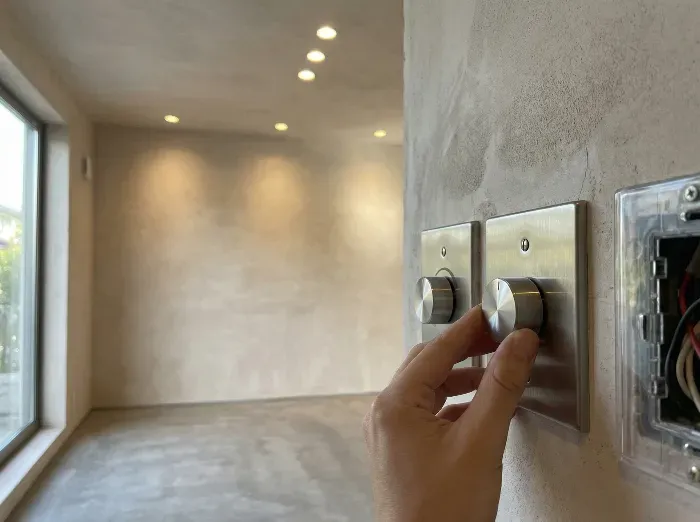 A hand adjusts a sleek metallic dimmer switch on a textured wall in a minimalist room with recessed ceiling lights and a large window.