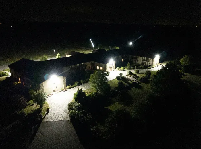 Aerial nighttime view of a lighted rural building complex with sloped roofs, surrounded by trees, driveway, and illuminated pathways in darkness.