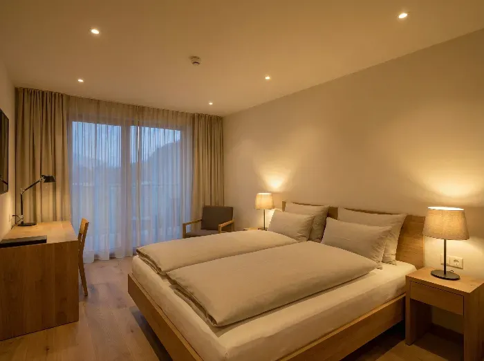 Spacious hotel bedroom with two single beds joined together, wooden desk and chair, large curtained windows overlooking misty mountains, and warm overhead lighting.
