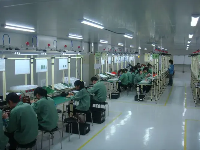 Workers in green uniforms assembling electronic components on long workbenches in a brightly lit, clean industrial factory environment.