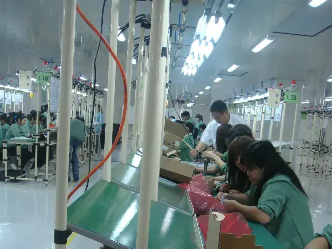 Group of workers in white uniforms assembling electronic components on green conveyor belts in a brightly lit modern manufacturing facility.