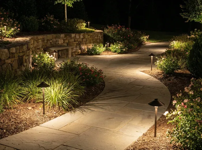 Curved stone pathway winds through lush nighttime garden, softly lit by ground-level path lights amid shrubs, flowers, and a stone wall.