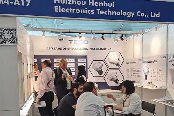 Trade show booth of Huizhou Hengnhui Electronics displaying LED lighting products, with professionals in business attire discussing innovations.