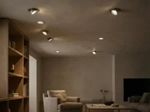 Contemporary room with beige walls, wooden bookshelf, beige armchair, small table with vase, and multiple recessed ceiling lights emitting warm glow.