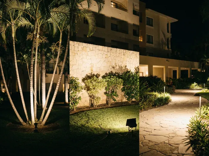 Nighttime scene of a luxurious tropical resort with palm trees illuminated by spotlights, stone walls, lush greenery, and a winding stone pathway lined with warm lights leading to a modern building.