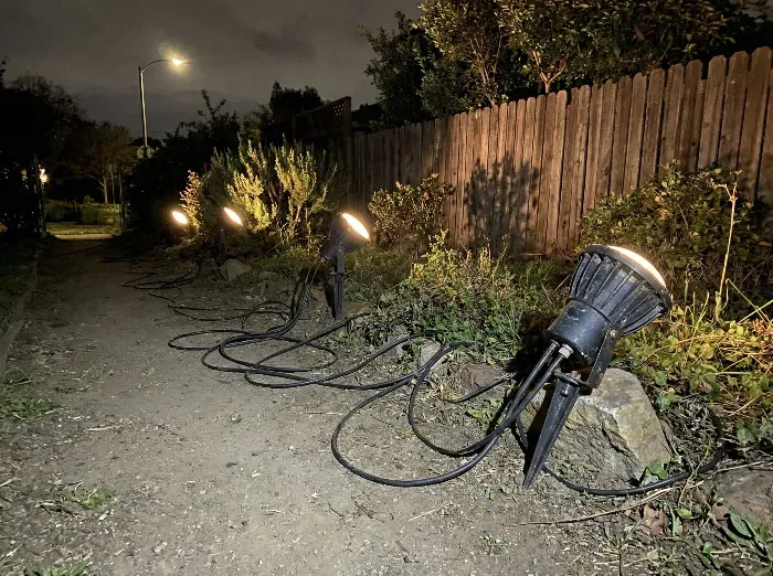 Several black cylindrical stake lights glow warmly along a dirt path at night, surrounded by bushes, rocks, and a wooden fence, with extension cords connecting them.