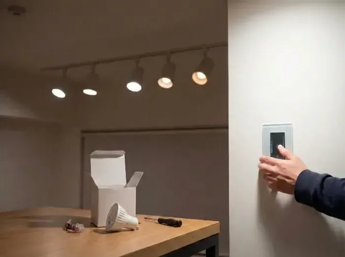 Split image: left side shows open box with white LED bulb, wires, and screwdriver on wooden table; right side depicts hand pressing wall switch beside illuminated track lights.