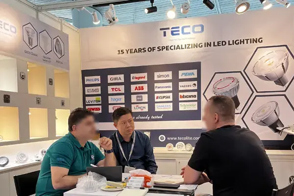 Trade show booth for TÉCO LED lighting company, displaying various LED bulbs and panels, partner brand logos like Philips and Osram, with professionals seated at a discussion table.