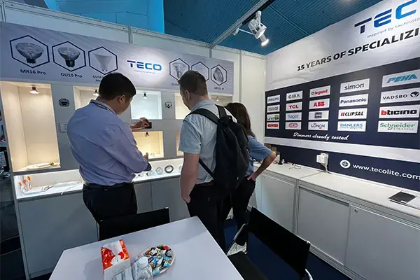 Trade show booth for TECO with visitors examining electrical displays, banners showing 18 years of specialization, and partner logos like Siemens and Schneider Electric.