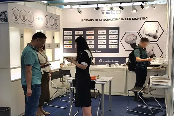 Trade show booth displaying Tecol LED lighting products, with banners celebrating 15 years, people browsing displays, and partner logos like Philips and Osram.
