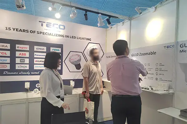 Trade show booth for TEGO displaying LED lighting products, company logos, and banners celebrating 15 years of specialization, with professionals viewing zoomable lamps and fixtures.