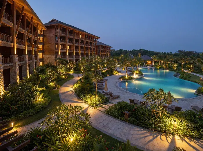 Luxurious wooden resort buildings surround a lit curved swimming pool amid lush palm trees and gardens under twilight sky.