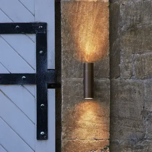 White wooden door with black iron hinges mounted on a rough stone wall, lit by a slim bronze cylindrical fixture emitting warm upward and downward light.