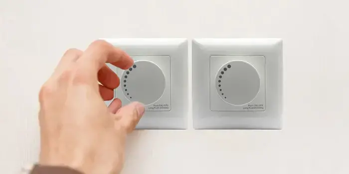 A person's hand gently turns the circular knob on a white wall-mounted dimmer switch, positioned next to an identical unused switch on a plain background.