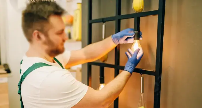 Bearded man in a white shirt, green suspenders, and blue gloves holding a glowing incandescent bulb in front of a black metal rack with hanging lights and yellow décor.