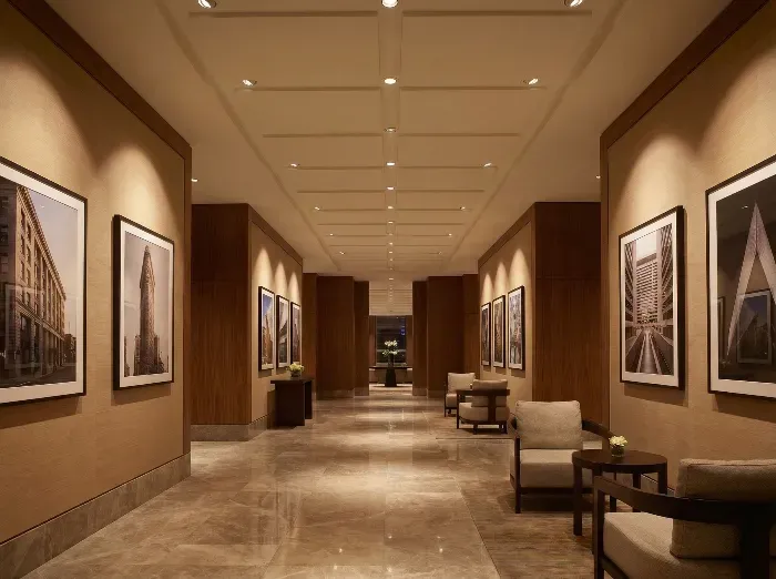 Long, warmly lit hallway with wooden panels, beige walls, large framed photos of modern buildings, and armchairs near the end.