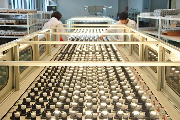 Two workers in white lab coats inspect rows of small black and white LED bulbs on a conveyor belt in a brightly lit factory setting with organized component shelves.