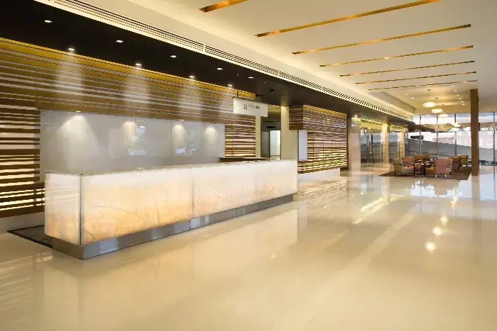 Spacious luxury hotel lobby with a sleek white onyx reception desk, backlit wooden wall panels, warm ambient lighting, and comfortable lounge seating on polished marble floors.