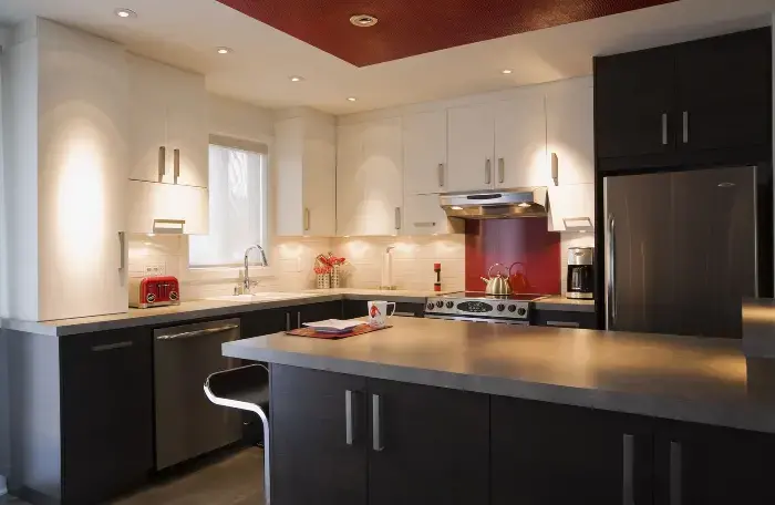 Bright modern kitchen with white upper cabinets, dark lower cabinets, red backsplash behind stainless steel stove, central island, appliances including toaster, kettle, and coffee maker.