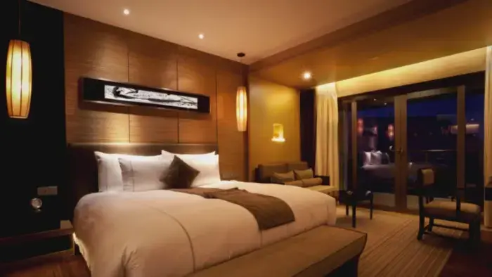 Elegant hotel bedroom with king-sized white bed, wooden panel walls, warm amber lighting from recessed and pendant lamps, and sliding glass doors to nighttime balcony.