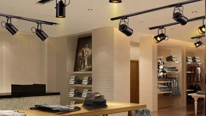 Interior of a contemporary clothing store with black ceiling-mounted track lights and spotlights illuminating wooden racks of folded shirts, hanging garments, and a front counter.