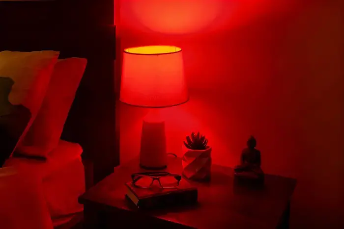 Cozy bedside table under red ambient lighting, holding a succulent in a geometric pot, small Buddha statue, eyeglasses, book, and candle holder beside a bed with pillows.