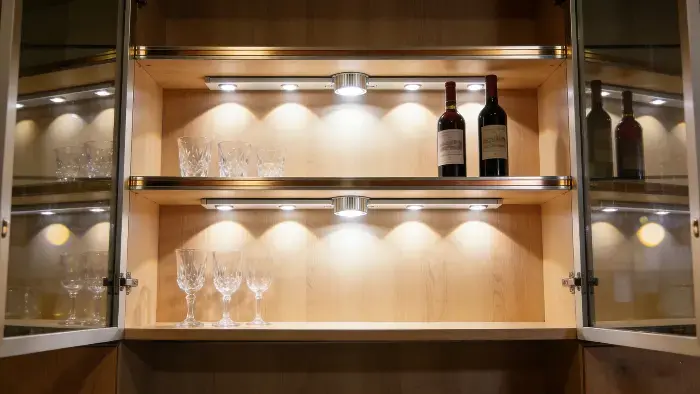 Open wooden cabinet with glass doors, featuring crystal wine glasses and red wine bottles illuminated by soft LED lights on shelves.