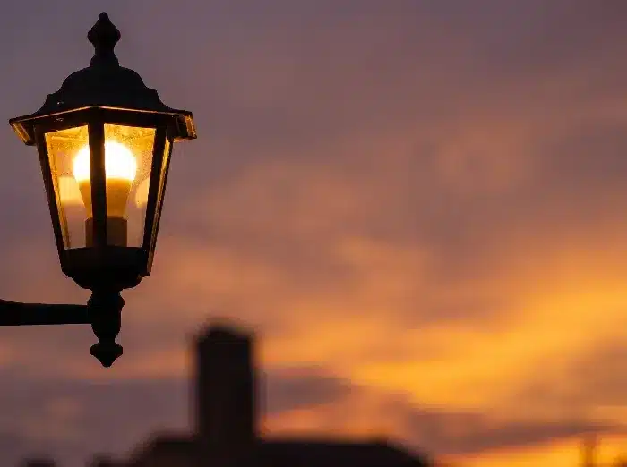 A lit vintage street lamp glowing against a vibrant orange and purple sunset sky with blurred buildings.