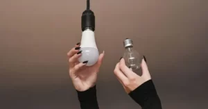 Hands with black nail polish holding a white LED bulb and a clear traditional bulb against a neutral background.