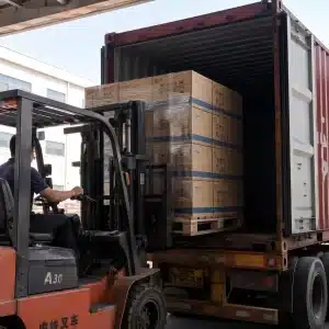 A worker operating an orange forklift to load stacked boxes into a red shipping container on a truck.